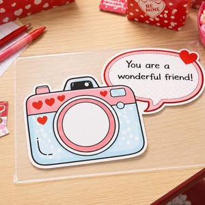 May include: A flat, rectangular card featuring a cartoon camera in light blue and pink with red heart accents. A speech bubble attached to the camera contains the text, "You are a wonderful friend!" The card is encased in a clear plastic sleeve.