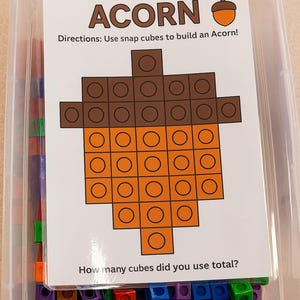 May include: A clear plastic container filled with colorful snap cubes and a card. The card features a pixelated acorn design made of brown and orange squares, with the word "ACORN" at the top and instructions below.