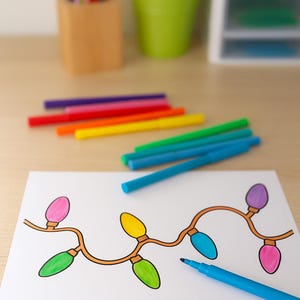 Puede incluir: Un dibujo de luces navideñas de colores sobre papel blanco, con un rotulador azul cerca. Varios rotuladores de colores están colocados encima del dibujo. Un portalápices de madera y un cubo verde están en el fondo.