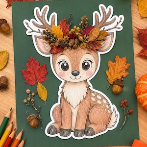 May include: A paper cutout of a brown fawn with large eyes and white spots, wearing a crown of dried autumn leaves, acorns, and berries. The fawn is centred on a dark green rectangular background, surrounded by loose fall leaves and acorns.