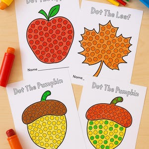 May include: Four dot-to-dot activity sheets featuring an apple, a leaf, and two pumpkins. Each image has a dotted pattern for connecting. The sheets are on a light wood surface, with crayons and felt-tip pens nearby.