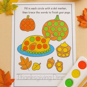 May include: Thanksgiving activity sheet featuring pumpkins, turkey, and acorns. The sheet instructs to fill circles with a dot marker and trace words. A dot marker, watercolour paint set, and autumn leaves are also visible.