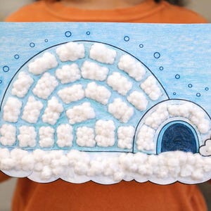 May include: A handmade craft project depicting an igloo. The igloo is drawn on blue paper and decorated with cotton balls to represent snow. The artwork includes a rounded entrance and a cloudy sky with blue circles.