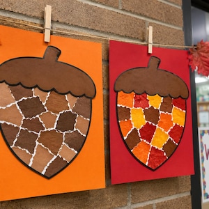 Puede incluir: Dos manualidades de papel con forma de bellota colgadas de una cuerda con pinzas de madera. Una bellota está sobre papel naranja con piezas de mosaico marrón, y la otra sobre papel rojo con piezas de mosaico amarillo, naranja y marrón.