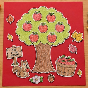 Puede incluir: Un proyecto de manualidades en papel sobre fondo rojo con un árbol central de tronco marrón y follaje verde claro con siete manzanas rojas. Una ardilla sostiene una manzana junto a un cartel que dice PICK OF THE SEASON y una cesta de manzanas.