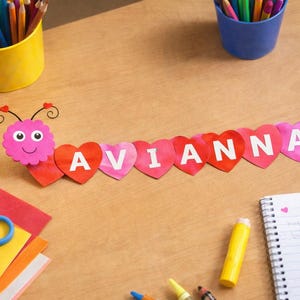 May include: A handmade name banner spelling "AVIANNA" with heart-shaped letters. The banner features a pink, smiling caterpillar head. Craft supplies, including colored pencils, scissors, and paper, are arranged on a wooden surface.