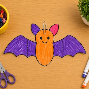 Puede incluir: Un recorte de papel de un murciélago con cuerpo naranja y alas moradas, con un clip plateado sujeto en la parte superior. El murciélago tiene dos pequeños ojos negros, una boca negra curvada y el interior de las orejas de color rosa.