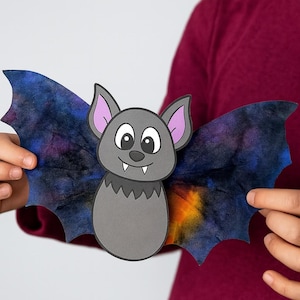 Puede incluir: Un murciélago de papel gris con alas grandes, una cara sonriente y orejas con forro morado. Las alas son de color azul oscuro con patrones coloridos tipo acuarela. Una divertida decoración de Halloween hecha a mano.