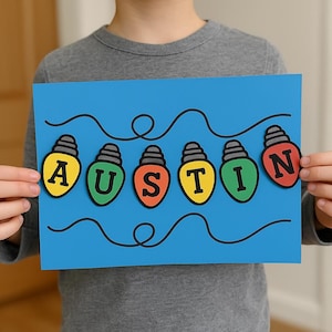 May include: A blue card displaying the name "AUSTIN" in the form of colourful light bulbs. Each bulb is a different colour, with the letters in black. The bulbs are linked by a black line, creating a festive design.