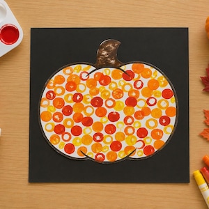 May include: A black square piece of paper featuring a pumpkin shape filled with circular stamps in shades of yellow, orange, and red. The pumpkin has a brown stem. Nearby are a paint palette, a bottle of school glue, three markers, and autumn leaves.