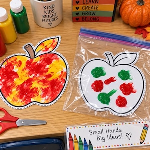 May include: Two apple-shaped paper cutouts, one painted with red and yellow swirls and one inside a clear plastic bag with red and green paint blobs. Includes red scissors, paint bottles, a mug with text, a sign reading Small Hands Big Ideas!