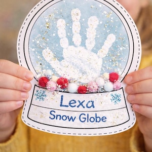 Può includere: Un artigianato di globo di neve fatto a mano con un'impronta di mano all'interno. Il globo di neve ha uno sfondo blu con glitter e pompon in basso. Il nome "Lexa" e "Snow Globe" sono scritti in basso.