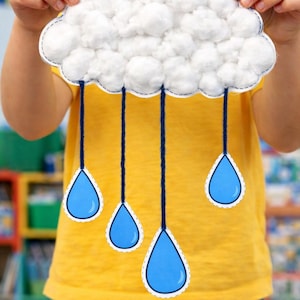 May include: A handmade cloud craft with cotton ball clouds and blue raindrop cutouts. The cloud is held up by a person wearing a yellow shirt and blue jeans. The craft is a fun, educational project.