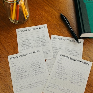 Può includere: Una raccolta di moduli "Avviso di riflessione sul comportamento", una penna, un taccuino verde e un barattolo di matite su una superficie di legno. I moduli hanno sezioni per nome, data e insegnante, con dettagli.