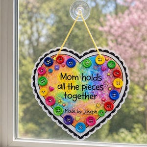 May include: A heart-shaped wall hanging with a rainbow-colored background and glitter. It features various colorful buttons attached to the surface and the text "Mom holds all the pieces together" and "Made by: Joseph" in black lettering.