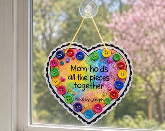 May include: A heart-shaped wall hanging with a rainbow-colored background and glitter. It features various colorful buttons attached to the surface and the text "Mom holds all the pieces together" and "Made by: Joseph" in black lettering.