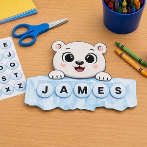 Może przedstawiać: Projekt z papieru przedstawiający rysunkową głowę niedźwiedzia polarnego na jasnoniebieskiej bryle lodu, z imieniem JAMES ułożonym z okrągłych wyciętych liter. Obok znajdują się arkusz naklejek z alfabetem, niebieskie nożyczki i klej w sztyfcie.