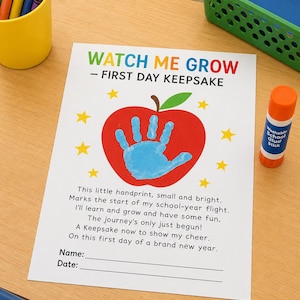 以下が含まれることがあります： WATCH ME GROW - FIRST DAY KEEPSAKEと書かれた白い紙。中央に赤いリンゴのグラフィックと青い手形があり、その周りを黄色い星が囲んでいる。テキストには詩が含まれ、下部には名前と日付を記入するための線がある。