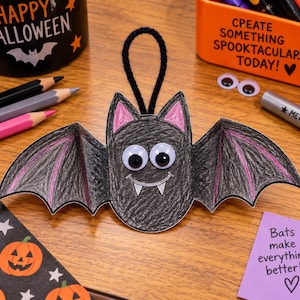 Puede incluir: Un adorno de murciélago de papel hecho a mano, coloreado en negro con detalles en rosa en las orejas y las alas. El murciélago presenta dos grandes ojos móviles, una boca sonriente dibujada con colmillos blancos y un cordón negro superior.