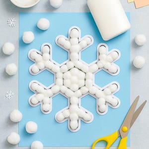 Può includere: Un progetto artigianale con un grande fiocco di neve fatto di batuffoli di cotone incollati su uno sfondo blu. Ulteriori batuffoli di cotone, una piccola ciotola, colla, forbici e carta sono disposti attorno al fiocco di neve. Le forbici hanno manici gialli.