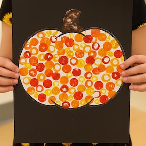 Puede incluir: Un papel negro con forma de calabaza recortada, lleno de círculos naranjas, amarillos y rojos. La calabaza tiene un tallo marrón. La persona lleva una camiseta negra con diseños de girasoles amarillos.