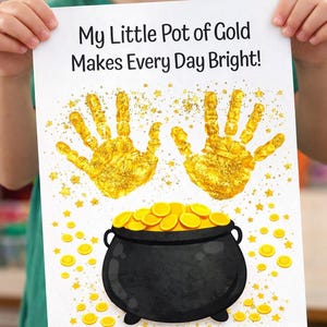 Puede incluir: Un papel blanco con el texto "My Little Pot of Gold Makes Every Day Bright!" presenta dos huellas de manos doradas, una olla negra llena de monedas de oro y estrellas doradas dispersas. La imagen es sostenida por una persona.