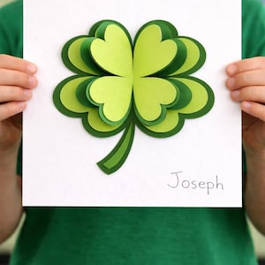 May include: A paper shamrock, crafted with layered green leaves, is displayed against a white backdrop. The name "Joseph" is written in cursive. The artwork is held by a person wearing a green top.
