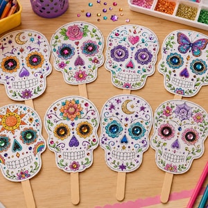 May include: Eight paper skull cutouts attached to wooden sticks, each decorated with colorful glitter, intricate floral patterns, and various adhesive gemstones. The skulls feature diverse designs including suns, moons, butterflies, and spiderwebs.