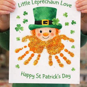 Pode incluir: Uma impressão artística do Dia de São Patrício com um design de duende. A imagem mostra um duende com um chapéu verde e impressões de mãos laranja como 'cabelo'. As palavras "Little Leprechaun Love" e "Happy St Patrick's Day" estão impressas.