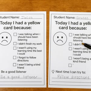 Può includere: Due schede bianche stampate con il testo "Oggi ho avuto un cartellino giallo perché:" e una faccia triste. Una scheda è compilata con il nome "Jacob" e l'altra con "Dralee". Una penna è appoggiata sulla seconda scheda. Le schede sono su una superficie di legno.