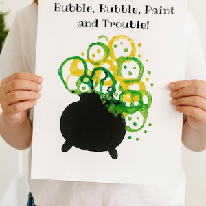 Puede incluir: Una hoja de papel blanca con el texto "Bubble, Bubble, Paint and Trouble!" se sostiene. La obra de arte presenta un caldero negro con burbujas verdes y amarillas que se elevan. La obra de arte es probablemente para Halloween.
