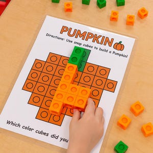 May include: A child's hand assembling a pumpkin from orange and green snap cubes. The image includes a printed guide with the word "PUMPKIN" and instructions. Scattered cubes are on a light wood surface.