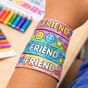 May include: A stack of colourful friendship bracelets with the word "FRIEND" in a playful font. Each bracelet features glitter and decorative elements like hearts, stars, and smiley faces. The bracelets are worn on a wrist.