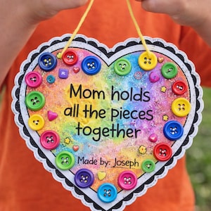 May include: A heart-shaped sign with a rainbow background and the text "Mom holds all the pieces together." The sign is decorated with colorful buttons and a scalloped edge. The sign is held by a person.