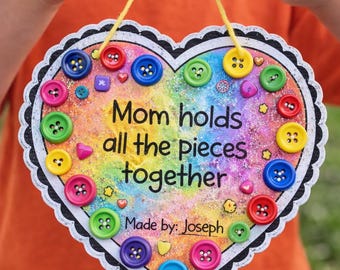 May include: A heart-shaped sign with a rainbow background and the text "Mom holds all the pieces together." The sign is decorated with colorful buttons and a scalloped edge. The sign is held by a person.