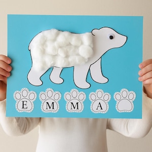 Könnte beinhalten: Ein handgefertigtes Eisbär-Bastelprojekt auf blauem Hintergrund. Der weiße Bär hat Watte als Fell und ein Wackelauge. Der Name "EMMA" ist in Pfotenabdruck-Ausschnitten geschrieben, was es zu einem personalisierten Kunstprojekt macht.