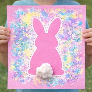 Könnte beinhalten: Rosa Osterei-Hasenkunst mit weißem, gepunktetem Umriss und einem flauschigen weißen Baumwollschwanz. Der Hintergrund ist mit bunten Pastellfarbspritzern verziert. Das Kunstwerk wird von einer Person hochgehalten.