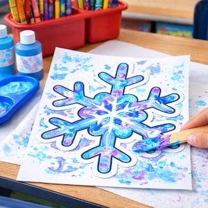 Puede incluir: Un proyecto de arte de copo de nieve con pintura azul y morada sobre papel blanco. La obra de arte está sobre un escritorio de madera, con botes de pintura, una paleta y crayones cerca. Una mano usa un palito de madera para pintar.