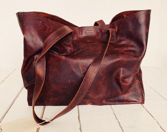 Puede incluir: Un bolso tote grande y abierto, hecho de cuero marrón oscuro desgastado. El bolso tiene dos correas largas para el hombro y una etiqueta adjunta. El cuero tiene un aspecto texturizado con variaciones de color, lo que sugiere un estilo vintage o rústico. El bolso está sobre una superficie de madera blanca.