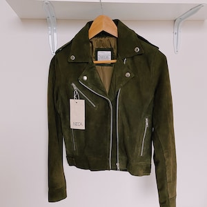 Puede incluir: Chaqueta de ante verde oliva de estilo motero clásico. Cuenta con cuello con solapas, cierre de cremallera asimétrico y múltiples cremalleras metálicas plateadas. La chaqueta cuelga de una percha de madera, con una etiqueta "NELA".