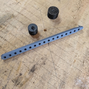 May include: A gray, plastic tool with evenly spaced holes and a pointed end, alongside two black, cylindrical objects with a central indentation. The items are arranged on a rough, wooden surface, suggesting a workshop or crafting context.