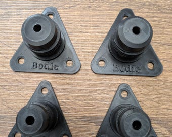 May include: Four black, triangular-shaped components with a cylindrical post and three mounting holes. The word "Bodie" is printed on each base. The components are arranged on a wooden surface.