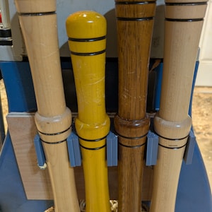 May include: Four wooden tool handles in varying colors: light wood, yellow, and dark brown. Each handle has a rounded top and is secured in a gray holder. The handles are for a woodworking tool.