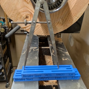 May include: A blue plastic ruler with the text "Divider Sizes" and markings for 50/75, 100/35, and 130. A metal divider tool is resting on the ruler.