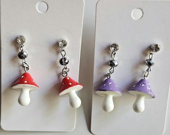May include: Two pairs of dangle earrings on white display cards. One pair features red mushroom charms with white spots, and the other pair features purple mushroom charms with white spots. Each earring has a clear crystal stud and a faceted bead.