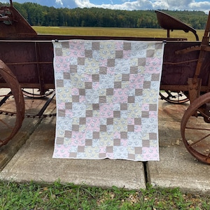 May include: A patchwork quilt with a checkered pattern in shades of pink, yellow, and brown. The quilt features a repeating pattern of small white sheep. The quilt is hanging on a rusty metal wagon.