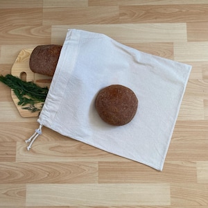 May include: A white cotton produce bag with two loaves of brown bread on a wooden chopping board with fresh dill.
