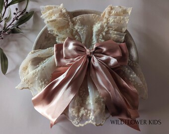 May include: A cream-colored dress featuring intricate floral embroidery and layered tulle ruffles on the shoulders. A large, dusty rose satin ribbon bow is centered on the front. The text "WILDFLOWER KIDS" is printed in the bottom right corner.
