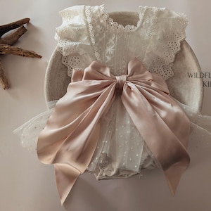 May include: A cream-colored romper featuring an eyelet lace bodice with ruffled sleeves, a large dusty pink satin bow at the waist, and a sheer tulle skirt overlay with white polka dots. The text WILDFLOWER KIDS is printed on the right side.