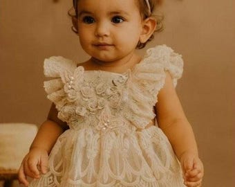May include: A cream-colored lace romper featuring ruffled sleeves, floral embroidery, and pearl accents, paired with a matching thin headband with a small lace bow. The garment has a scalloped hemline and a layered design with intricate detailing.